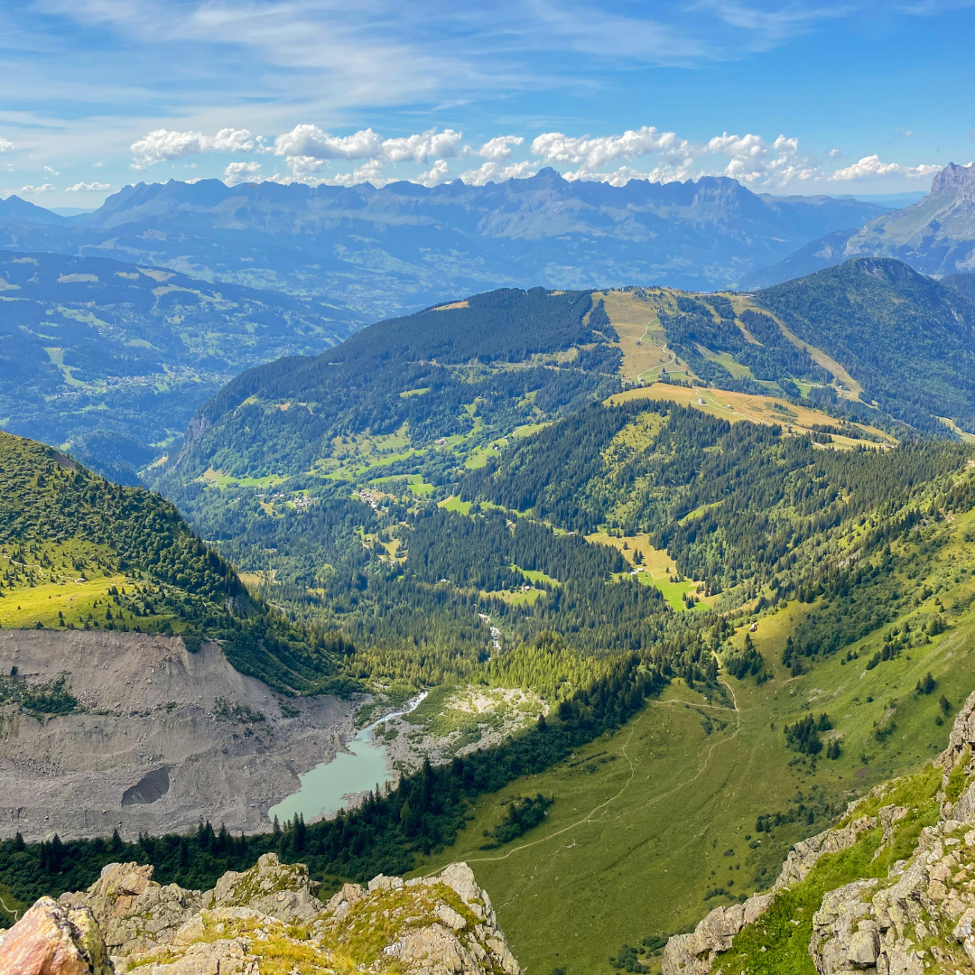 Green mountain with a lake at the bottom. Above is blue sky with scattered white clouds.