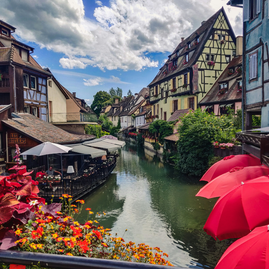 A view of a river below, taken from a bridge. The river is lined with buildings, a cafe on the left and some red umbrellas on the right.